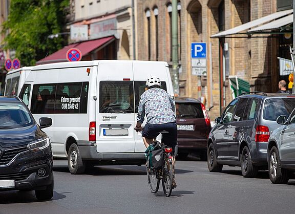 Radfahrer auf Geschäftsstraße neben parkenden Autos. 