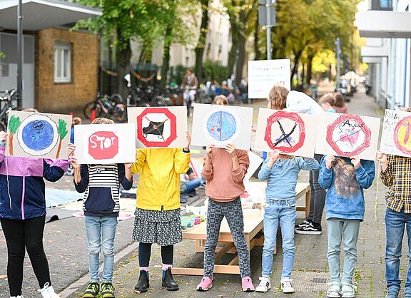 Kinder blockieren eine Straße, halten Plakate hoch und setzen sich damit für die Einrichtung von Schulstraßen ein.